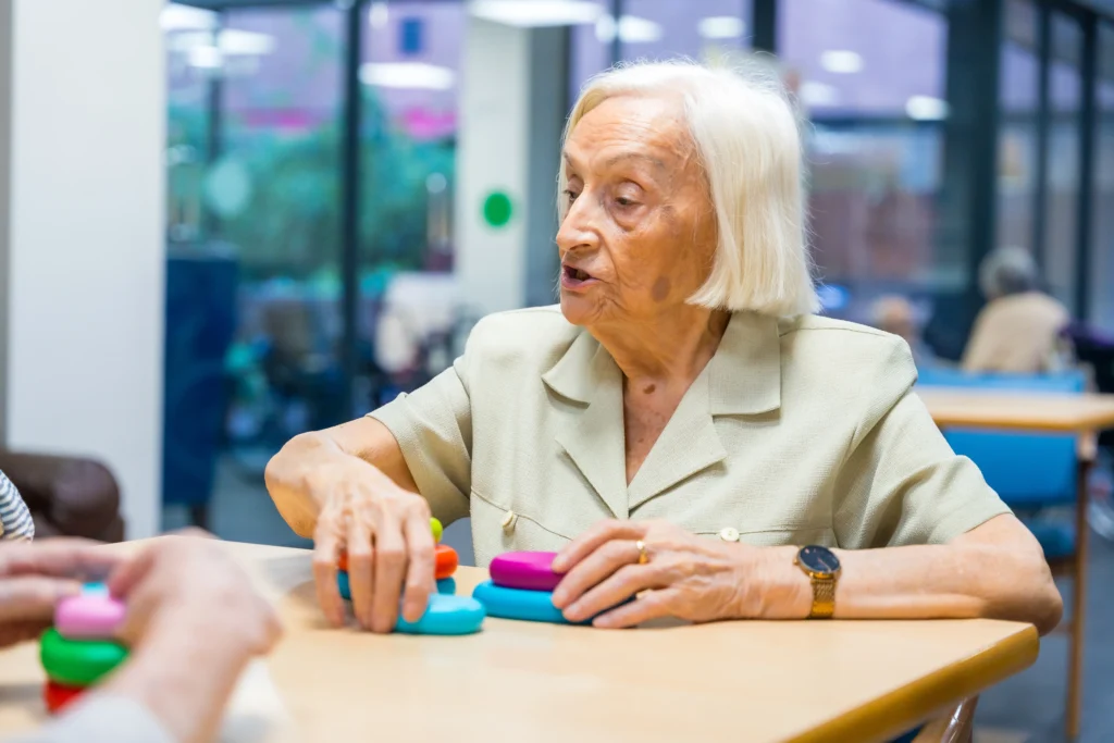 An elderly woman participating in an adult speech therapy or cognitive rehabilitation session using colorful stacking rings at a table.