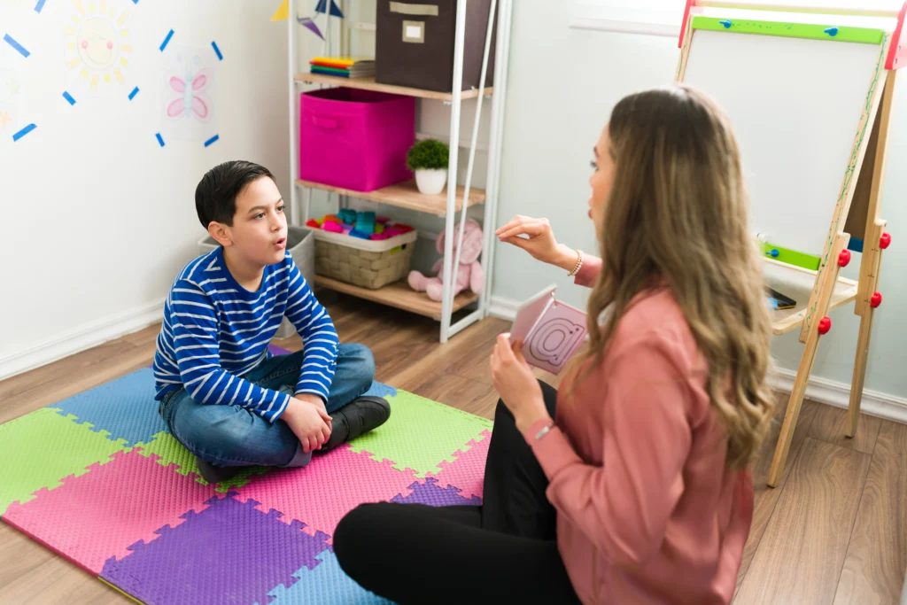 A pediatric speech-language pathologist sitting on a playroom floor with a young boy, using alphabet cards to teach letter sounds and speech articulation.