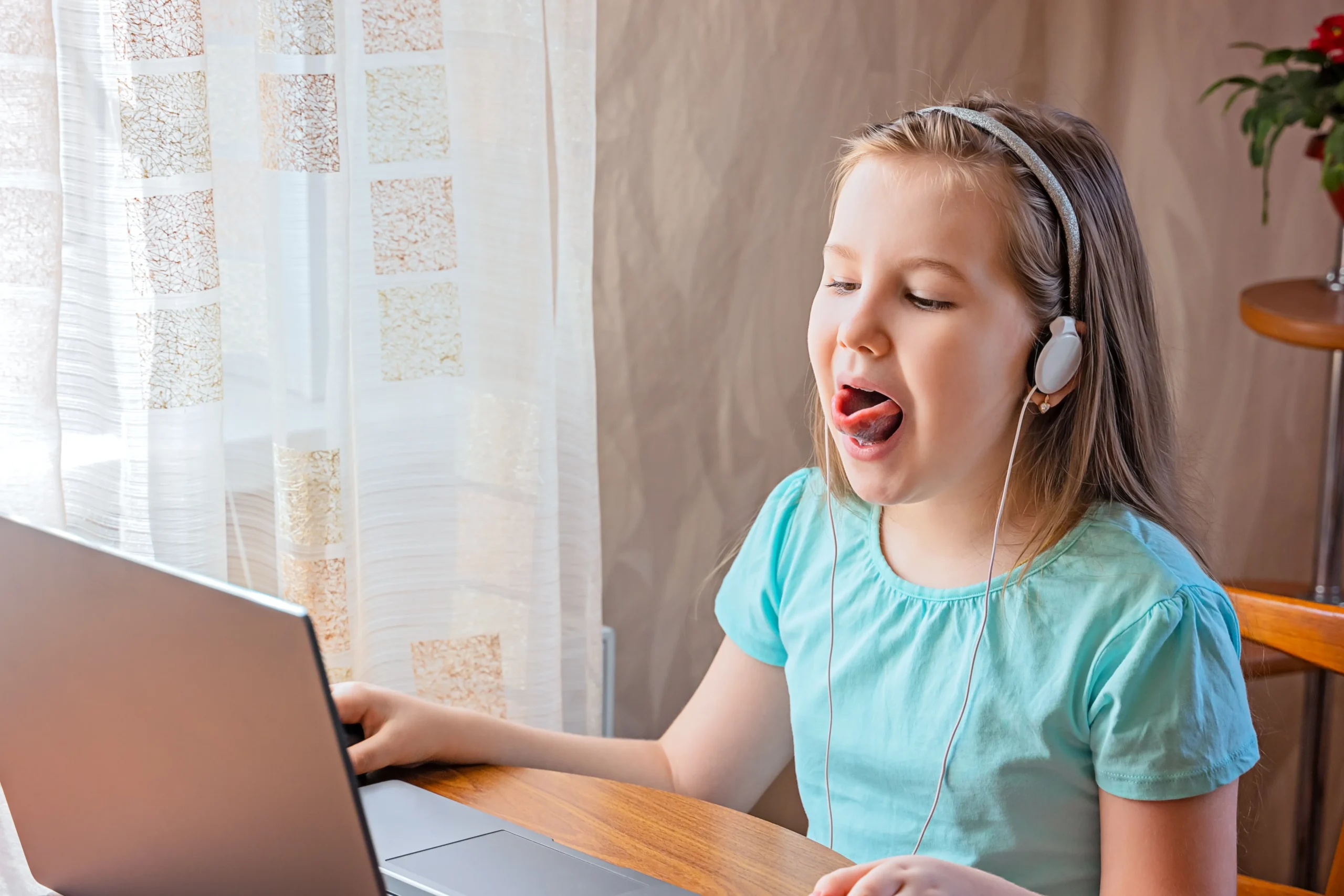 A young girl wearing headphones practicing tongue placement and pronunciation during a remote, online speech therapy session on a laptop.