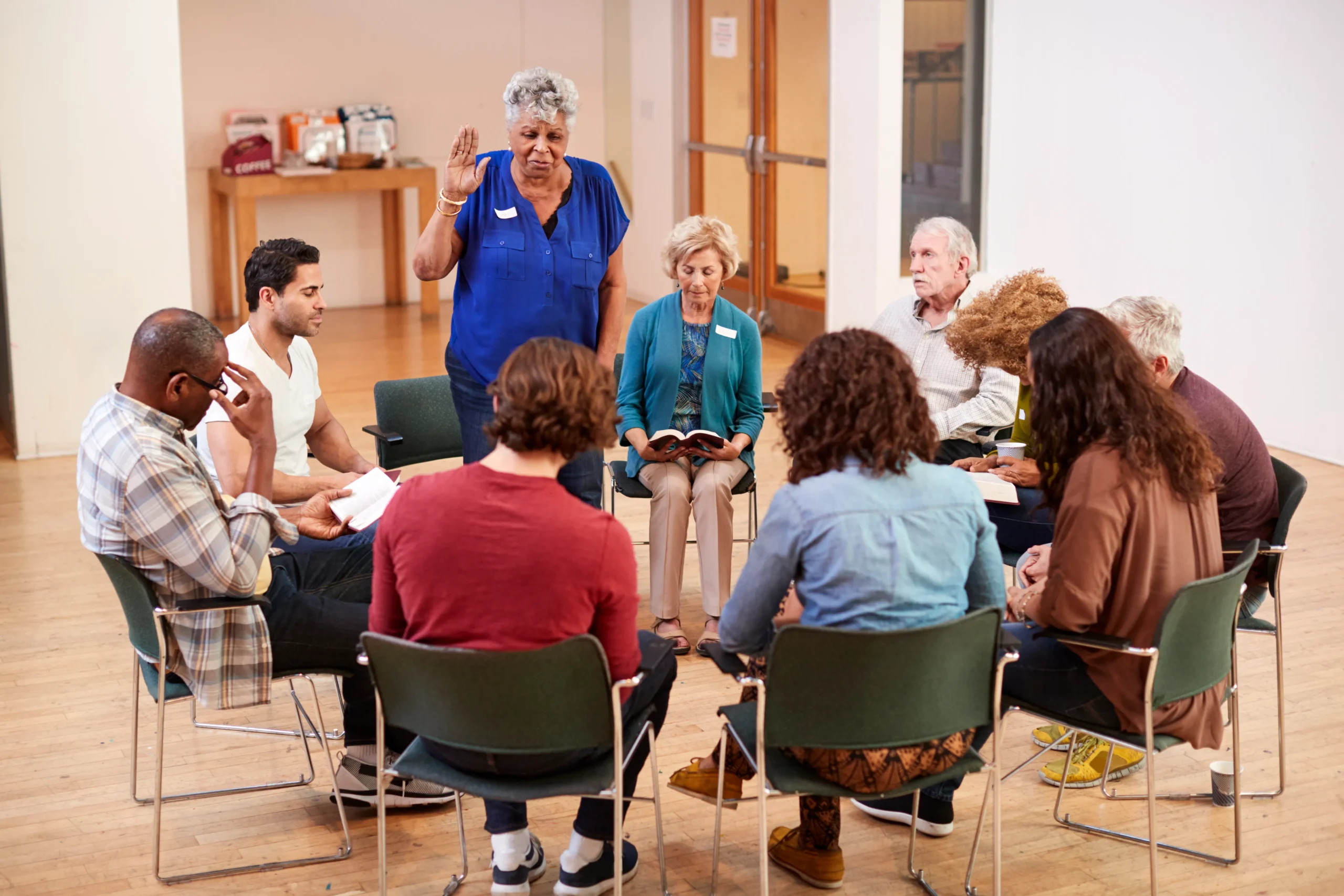 A diverse group of adults sitting in a circle during a community support group meeting or counseling session while a woman speaks.