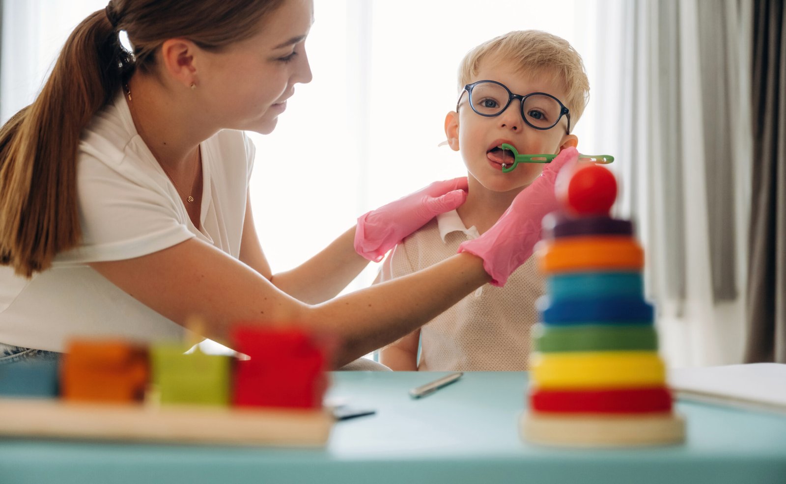 A female speech-language pathologist wearing pink gloves uses an oral placement therapy tool to assist a young boy with tongue positioning.