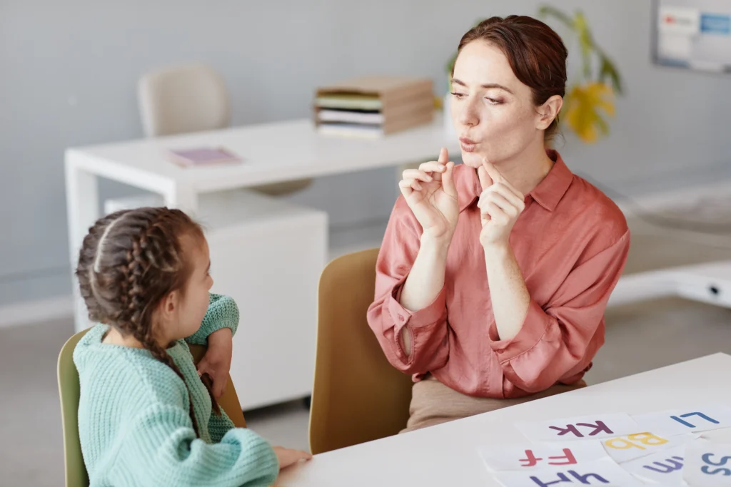 A speech-language pathologist demonstrating correct lip shape and mouth formation to a young girl during an articulation therapy session.