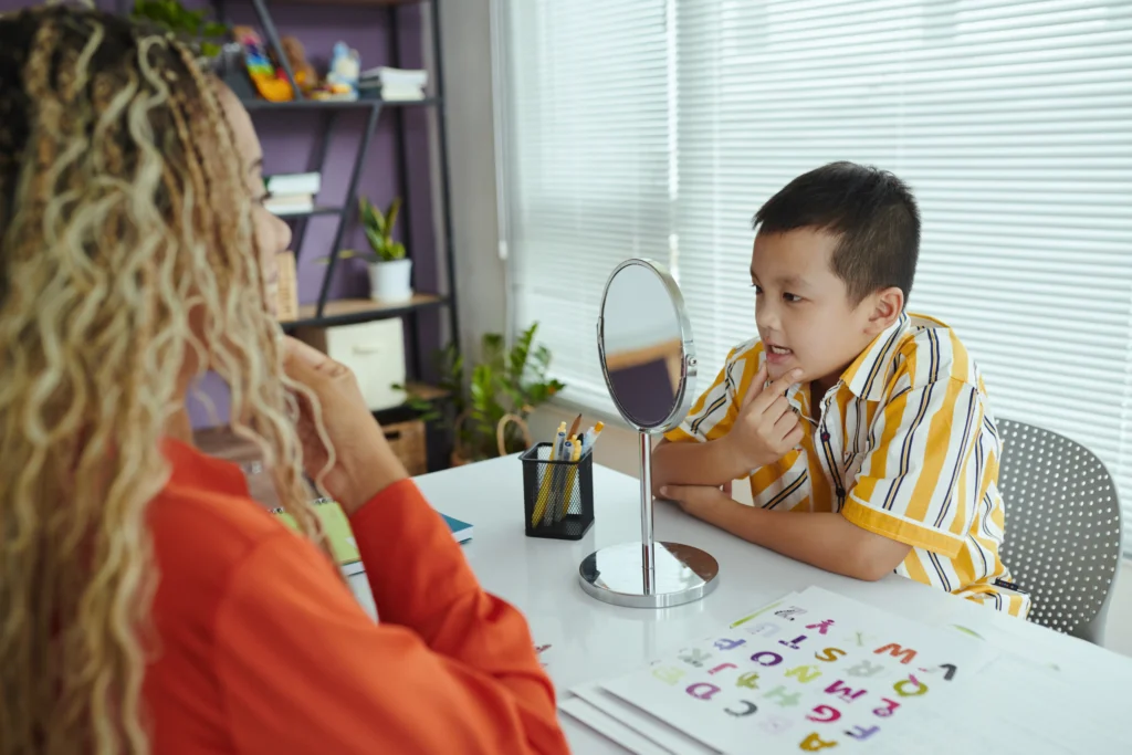 A young boy practicing mouth movements in a desk mirror during a pediatric speech therapy session with a female therapist.