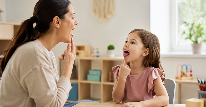 A speech therapist and a young girl face each other at a desk while practicing mouth positioning for target speech sounds.