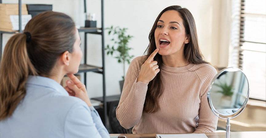 An adult woman practicing speech therapy with a professional, both pointing to their mouths next to a small vanity mirror.