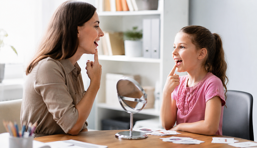 A speech therapist and a young girl sit at a table practicing speech placement, both pointing to their mouths while looking at each other, with a small mirror and picture cards on the table.