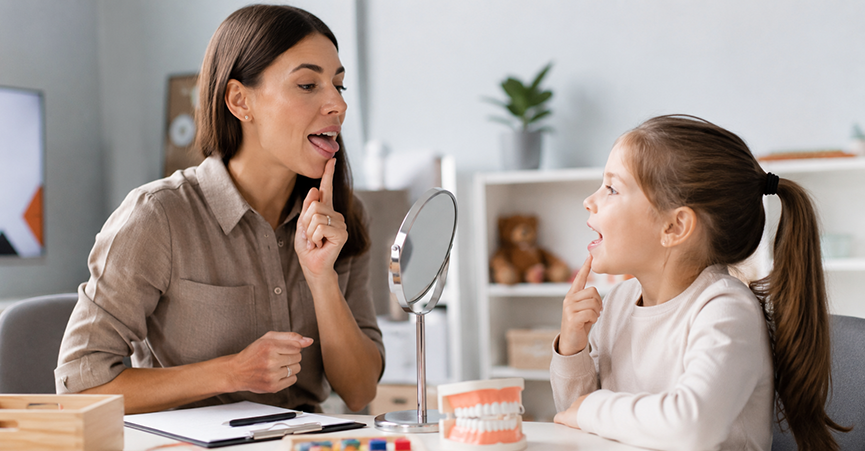 A speech therapist and a young girl sit across from each other at a desk, both pointing to their mouths during a therapy exercise, with a small mirror and a dental model on the table between them.